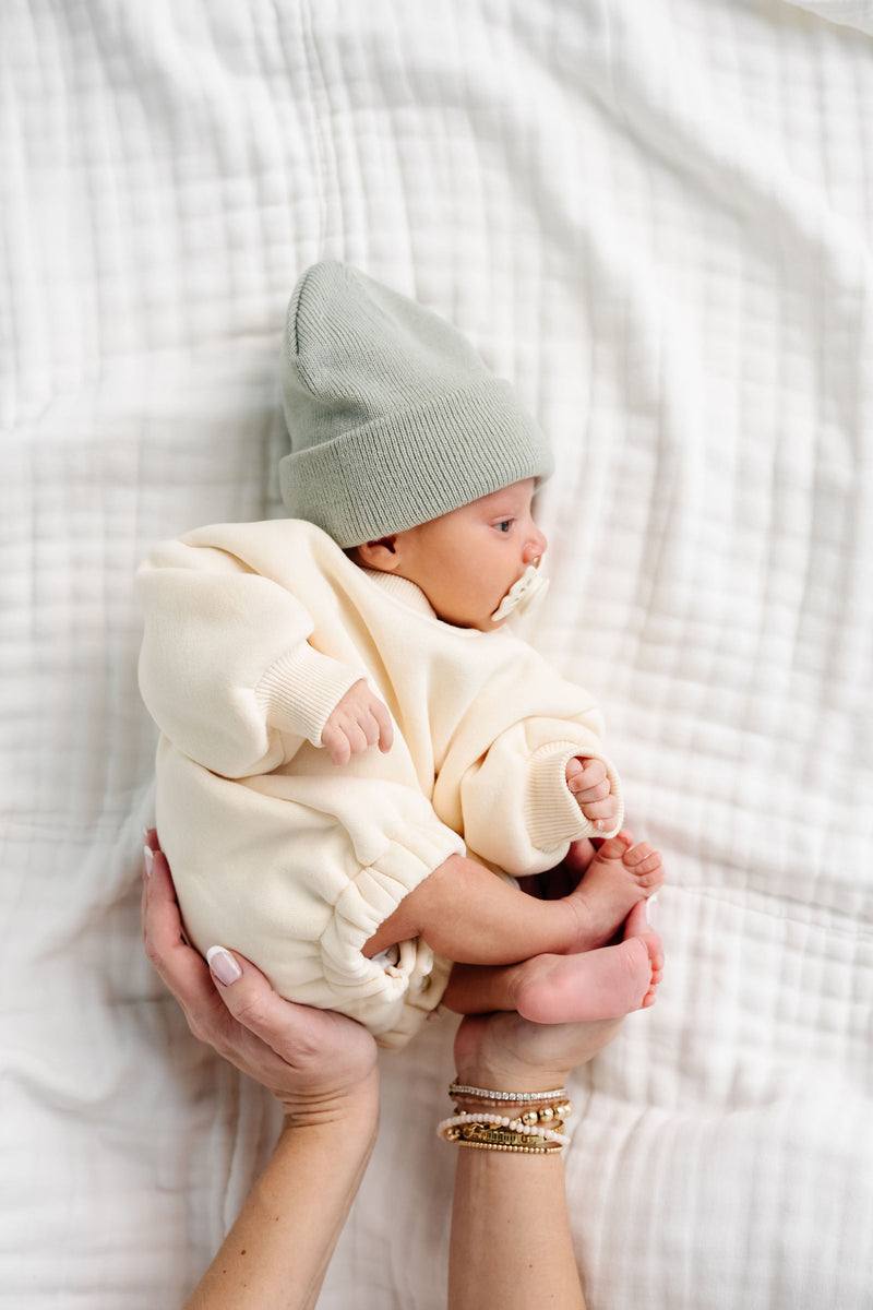 cream sweatshirt romper baby boy with beanie laying on bed